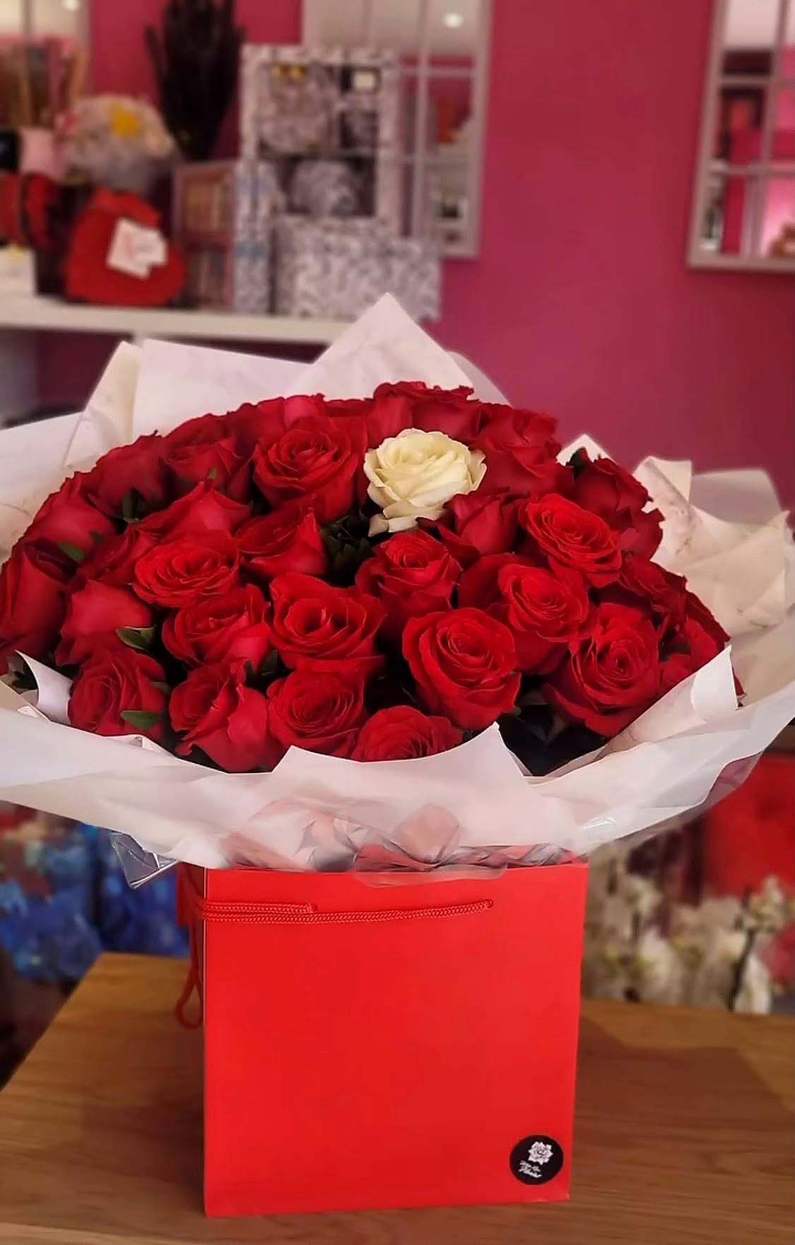 A large bouquet of red roses with one white rose in the centre, wrapped in white paper in a red box