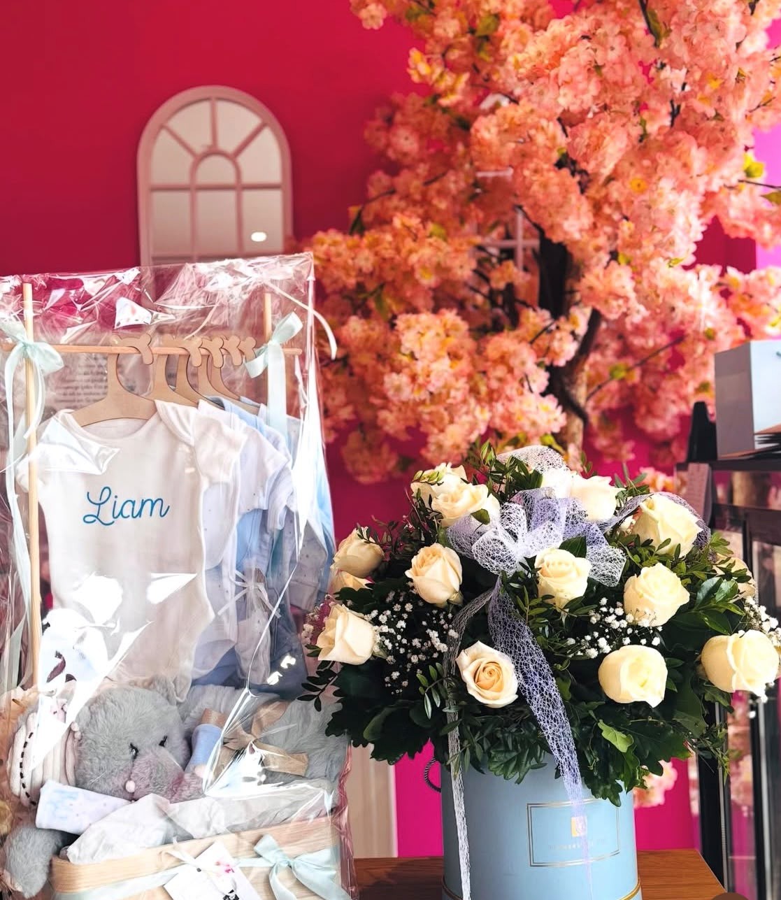 A baby hamper with clothes and a toy beside a hatbox of cream roses